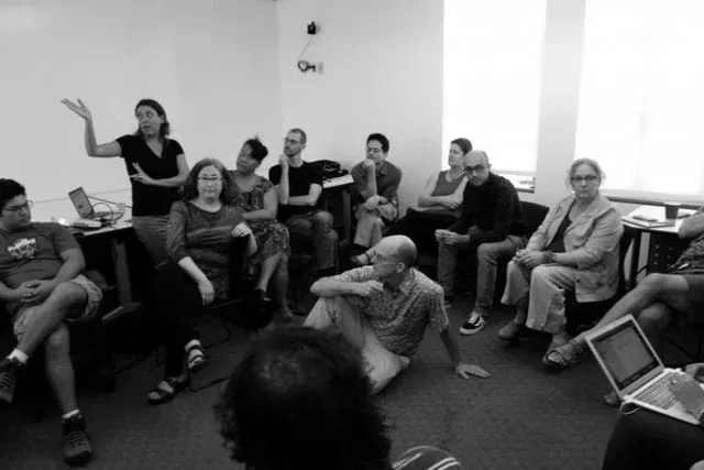 A facilitator gestures while addressing about ten participants seated in a loose circle on chairs and the floor of a small room, several with laptops open, during a workflow sprint discussion.