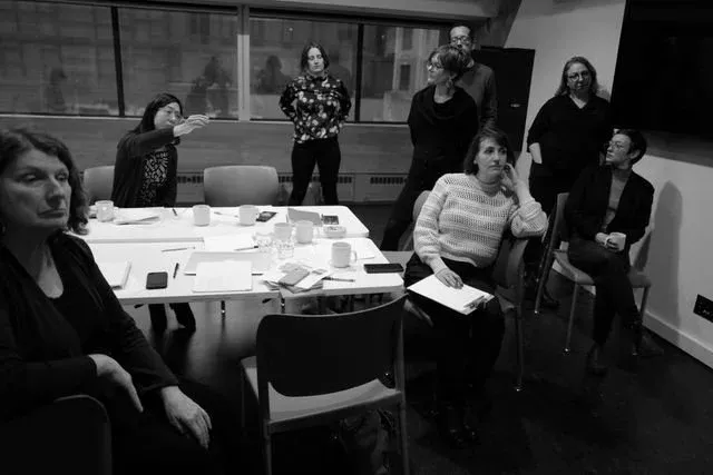 Evening sprint session in a meeting room: around ten participants sit and stand around a table strewn with notepads, papers and mugs, listening as one person gestures towards the far side of the room.