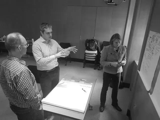 Workflow sprint in progress: a facilitator gestures mid-explanation to two colleagues standing around a blank flipchart on a whiteboard easel in a meeting room, with a second annotated flipchart sheet at the right.