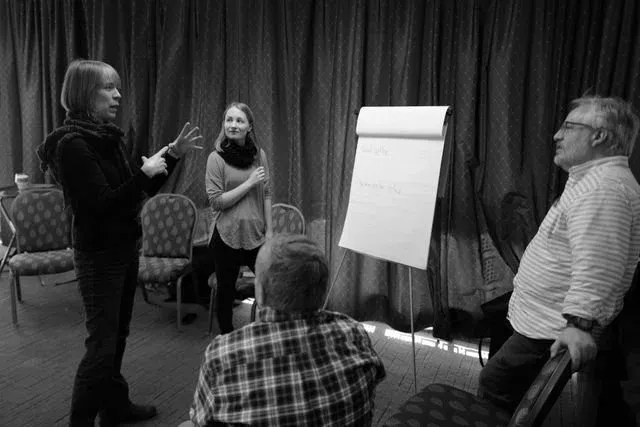 Group discussion during a workflow sprint: a participant gestures while explaining a point to three seated and standing colleagues beside a flipchart holding two short handwritten lines.