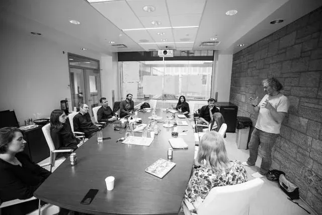 A facilitator stands against a stone wall presenting to roughly a dozen participants seated around a long boardroom table with papers, notebooks and drinks, in a room with a ceiling projector and glazed wall.